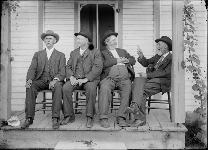 Four men in suits and hats are seated in chairs on the front porch