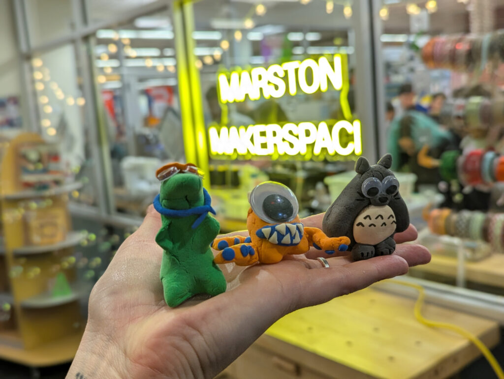 Three clay figures in front of neon sign that says "Marston Makerspace"