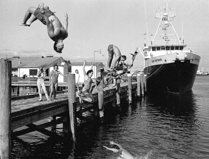 black and white photo of student back diving off a dock into the water. Other students on the dock with boat in background.